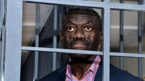 Reuters Kizza Besigye and a salmon pink and purple stripped shirt and a navy jacket seen behind bars