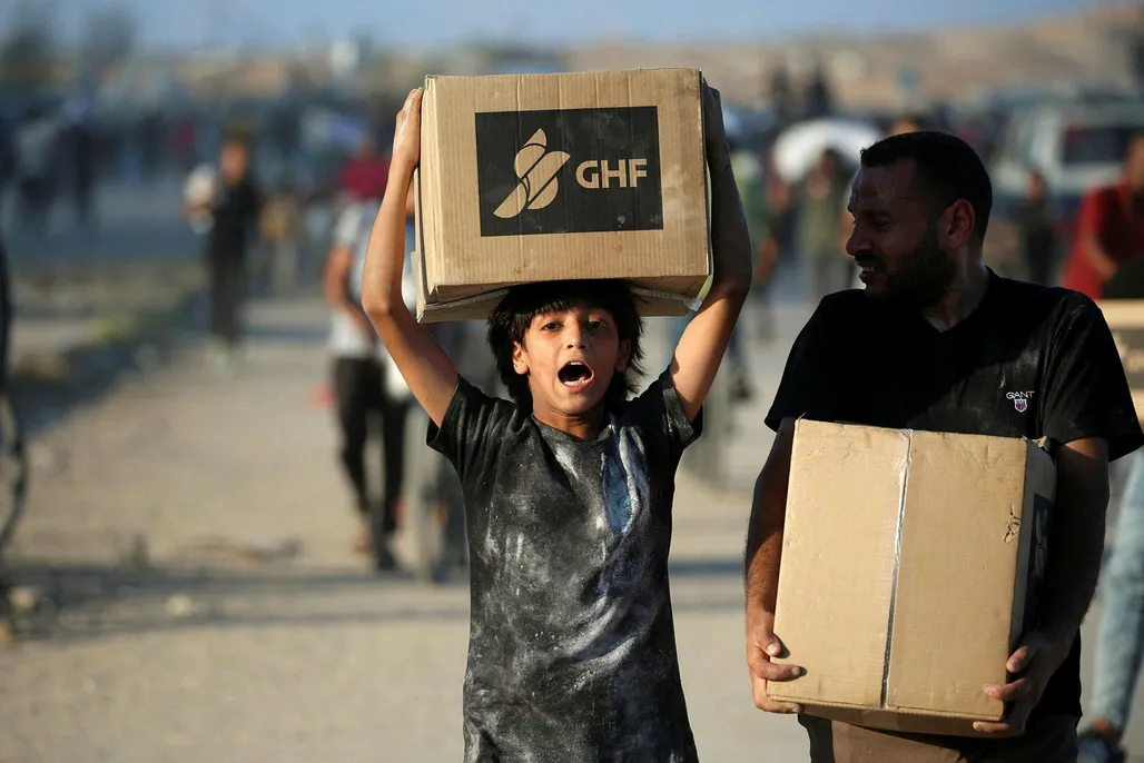 People carrying aid parcels, walk along the Salah al-Din road near the Nusseirat refugee camp in the northern Gaza Strip, used by food-seeking Palestinians to reach an aid distributution point set up by the privately-run Gaza Humanitarian Foundation (GHF), on June 25, 2025. (Photo by Eyad BABA / AFP)